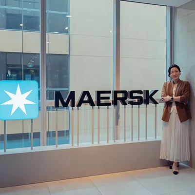 A woman stands beside a large illuminated Maersk logo inside a modern office with floor-to-ceiling windows.