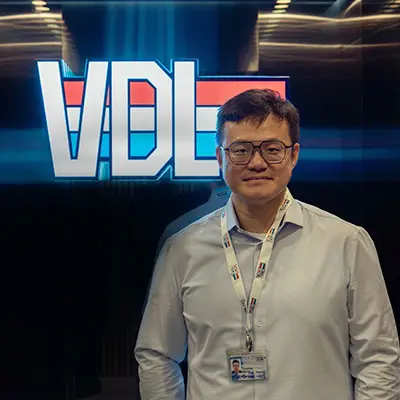 A man wearing glasses and a white shirt stands confidently in front of a brightly illuminated "VDL" logo. He is wearing an ID badge around his neck, suggesting a professional or corporate setting.