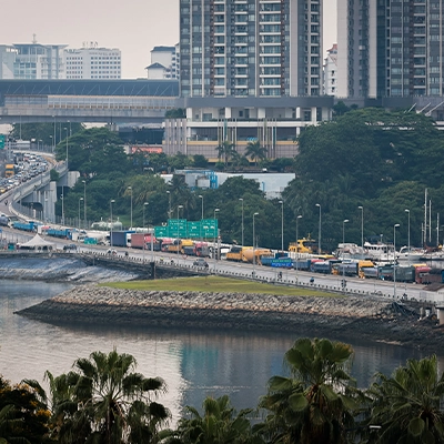 : A busy causeway connecting Singapore and Johor filled with cars and trucks, with city buildings and greenery in the background.
