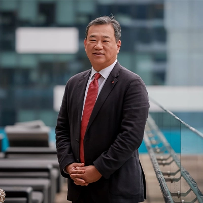 Lotte Retail Group vice-chairman and group CEO Sanghyun Kim smiling while standing outdoors on a balcony with modern glass buildings in the background.