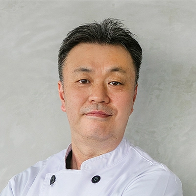 A confident man in a white chef's coat stands with arms crossed against a light grey background, looking directly at the camera with a slight smile.