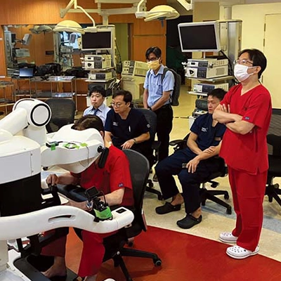 A group of medical professionals observes a demonstration of robotic surgical equipment in a hospital or clinical setting, with one individual actively operating the machine while others look on attentively.
