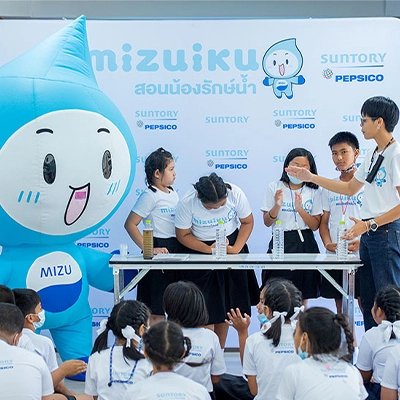 A group of children participates in an educational water conservation activity led by a presenter and accompanied by a large blue water droplet mascot named "Mizu" at a Suntory PepsiCo event. The setup includes a demonstration table and a backdrop with branding in Thai and English.